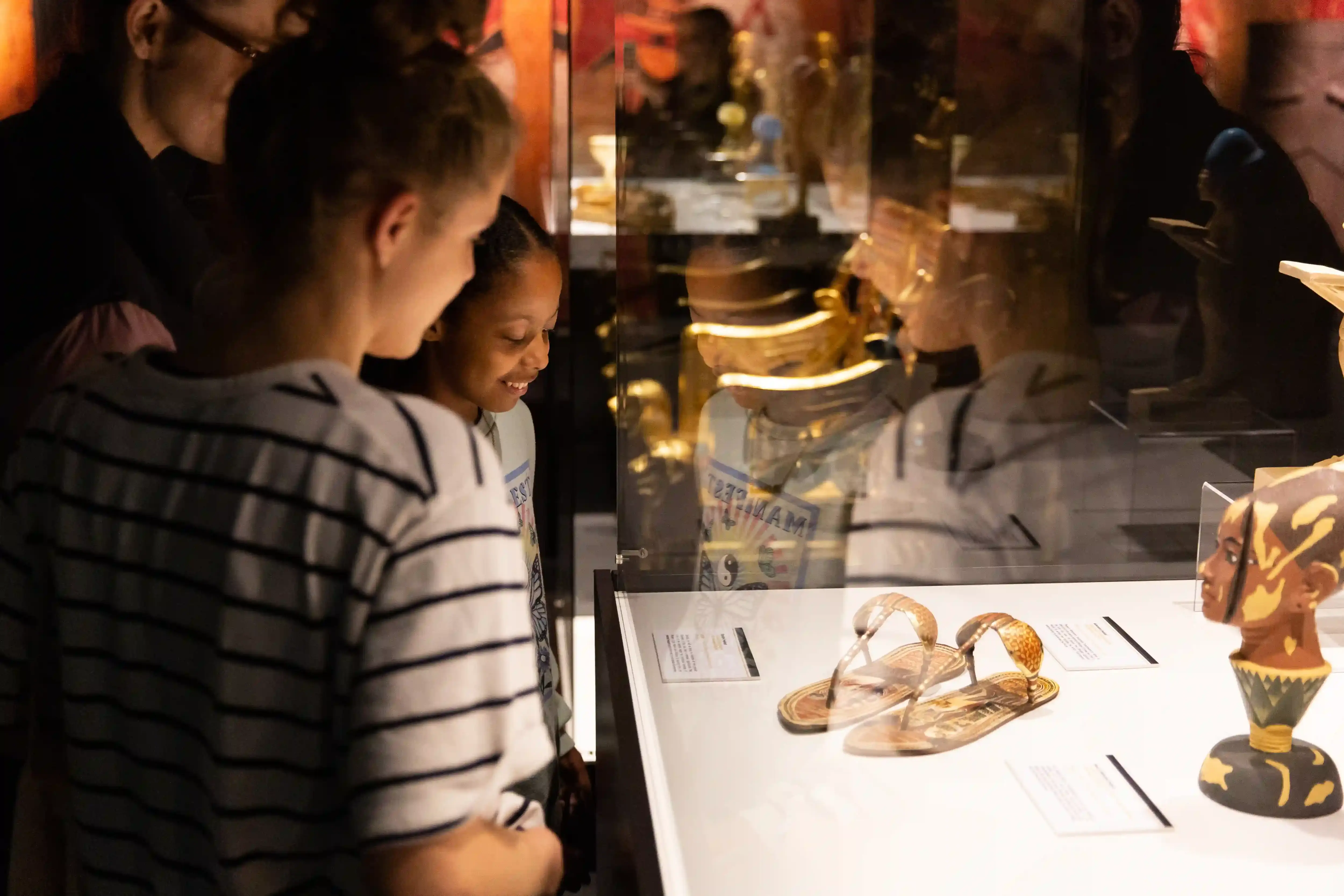 Tutankhamun-The-Immersive-Exhibition-Helsinki-1 aksi lasta katselee vitriinissä kultaisia sandaalijäljennöksiä ja lootuskukasta nousevaa päätä esittävää veistosjäljennöstä.