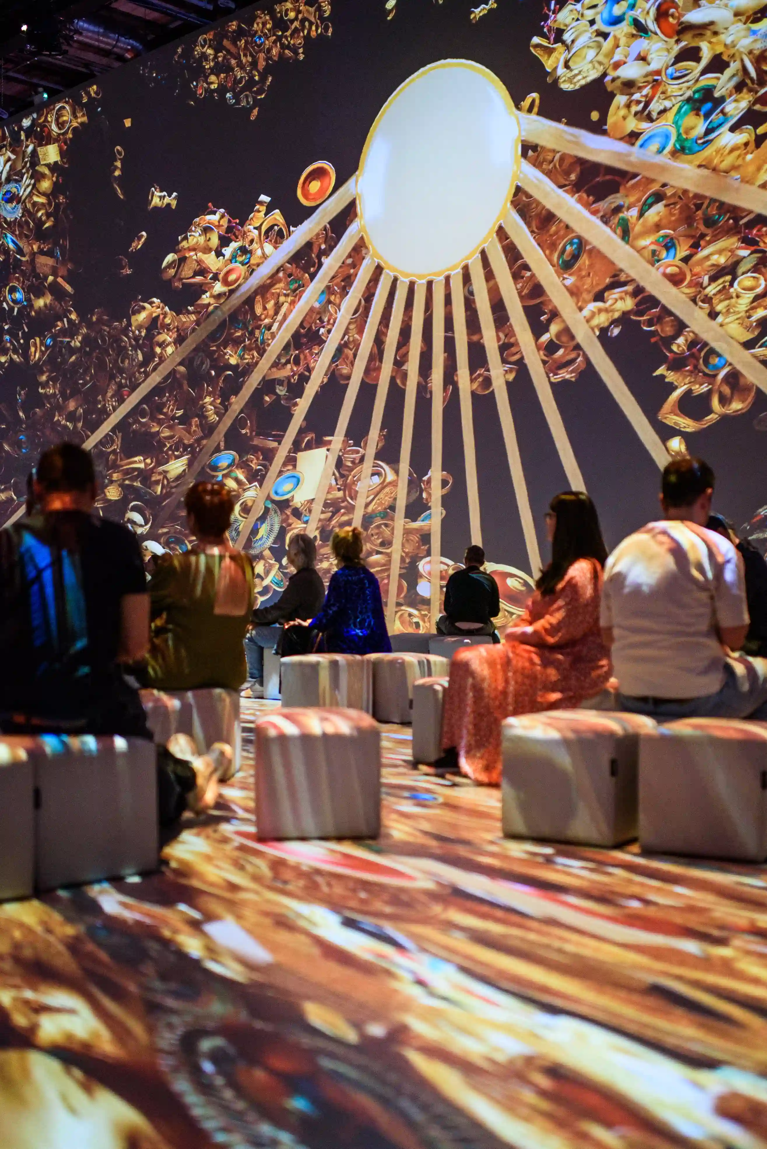 Tutankhamun-The-Immersive-Exhibition-Helsinki-3 Kävijät istuvat rahien päällä immersiivisessä salissa, jonka seinille ja lattialle heijastetaan kultaisia aarteita ja säteilevä aurinkokiekko.
