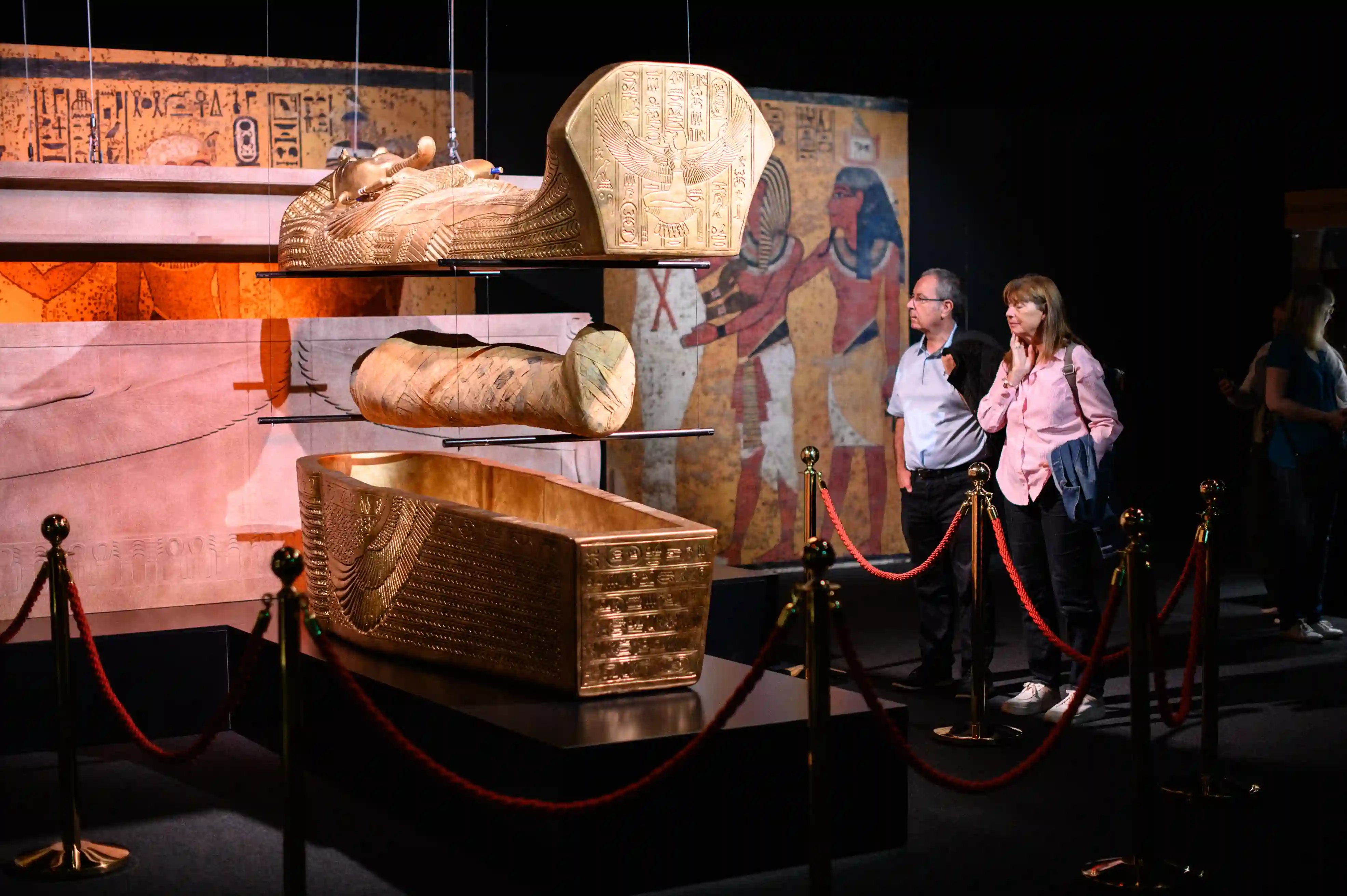 Tutankhamun-The-Immersive-Exhibition-Helsinki-4 Kävijäpari katselee Tutankhamonin kultaisen arkun kannen, muumion ja arkun pohjaosan jäljennöksiä hautamaalausten jäljennösten edessä.