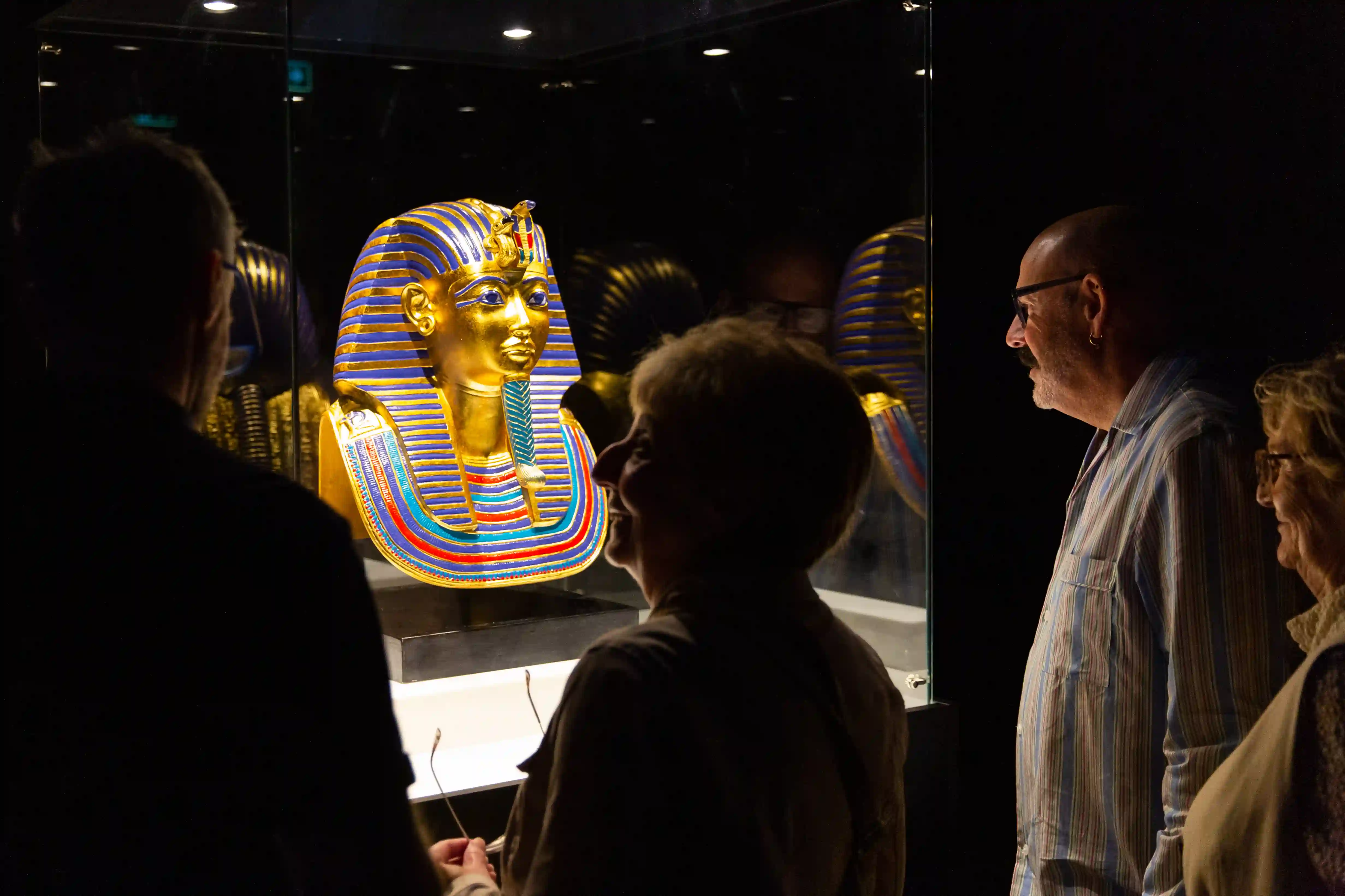 Tutankhamun-The-Immersive-Exhibition-Helsinki-8 Hymyilevät kävijät katselevat valaistussa lasivitriinissä olevaa Tutankhamonin kultaisen kuolinnaamion jäljennöstä hämärässä näyttelytilassa.