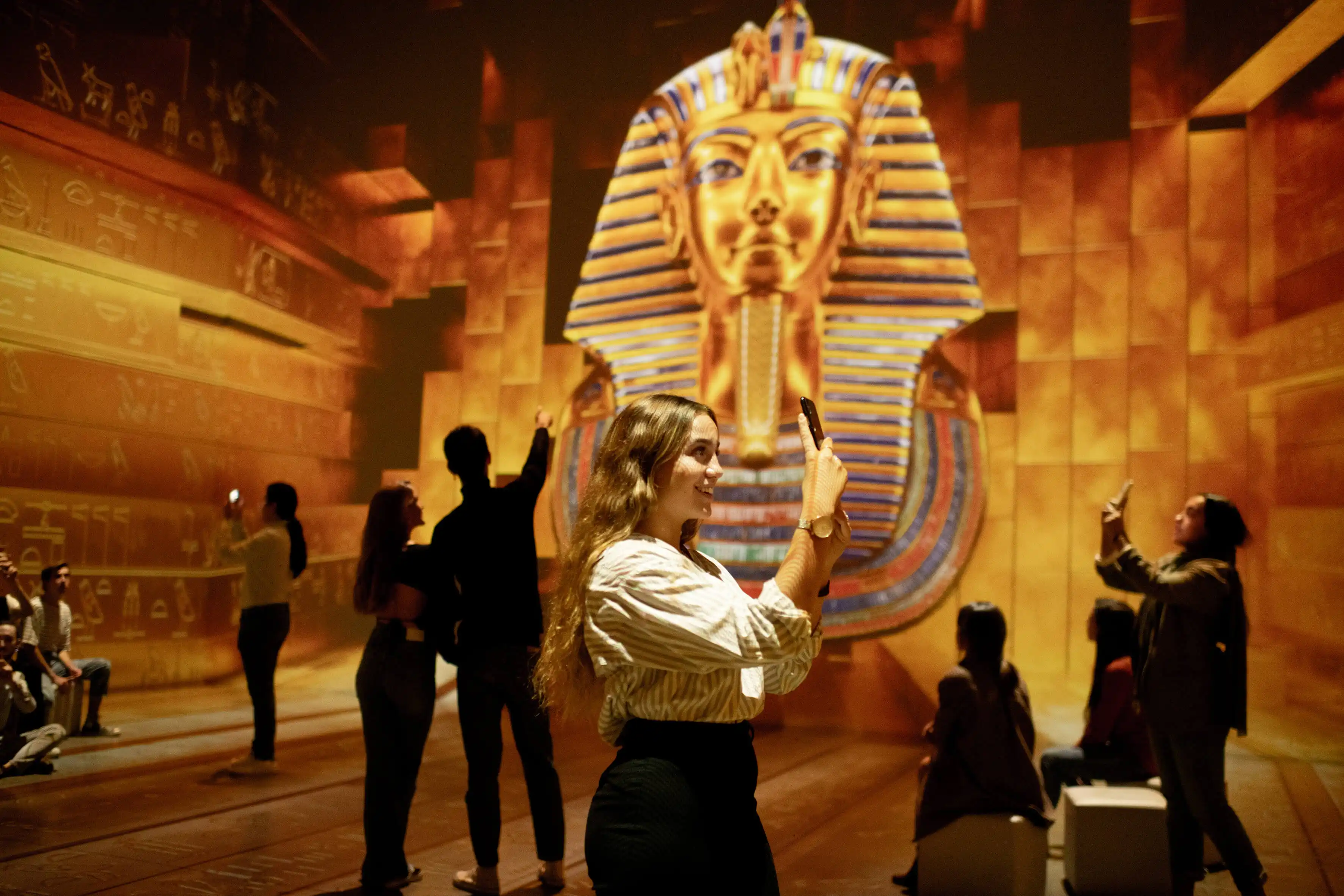 Kävijä ottamassa kuvaa suuren kultaisen Tutankhamon-naamion projektion edessä hieroglyfein koristellussa salissa.